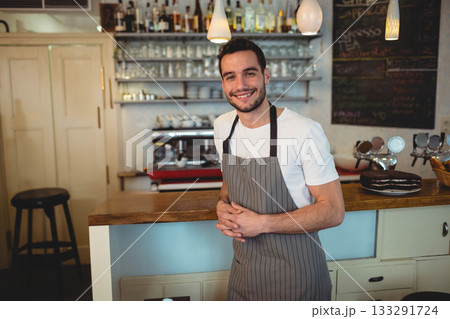 Male cafe worker wearing striped apron standing and smiling behind counter with espresso machine 133291724