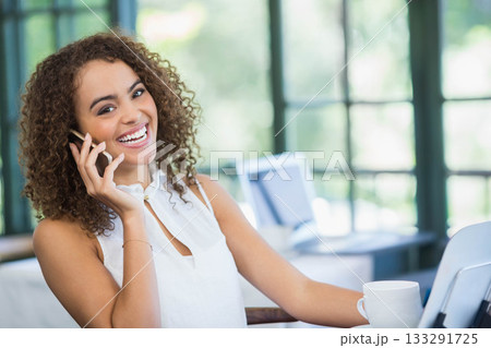 African American woman holding smartphone, smiling at desk in bright office with laptop, coffee mug African American woman holding smartphone, smiling at desk in bright office with laptop, coffee mug 133291725