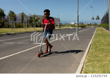 African American child boy riding silver scooter along coastal path beside road with backpack African American child boy riding silver scooter along coastal path beside road with backpack 133291734