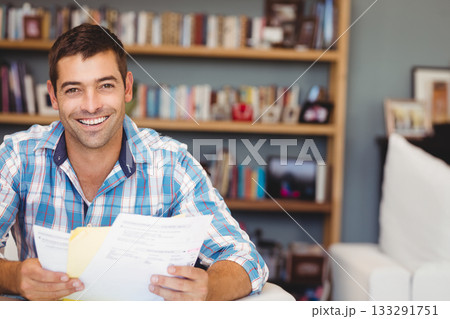 Man in plaid shirt holding printed documents, smiling on couch in home office, copy space 133291751