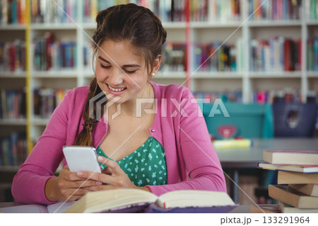 Teenage student smiling scrolling smartphone beside open book on library table bookshelves, bins Teenage student smiling scrolling smartphone beside open book on library table bookshelves, bins 133291964