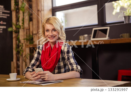 Woman sitting at wooden table in coffee shop, sipping coffee and checking smartphone with newspaper 133291997