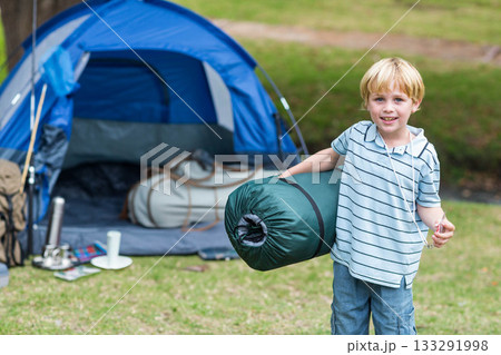 Boy carrying rolled sleeping bag while standing on grassy campsite by blue dome tent, copy space Boy carrying rolled sleeping bag while standing on grassy campsite by blue dome tent, copy space 133291998