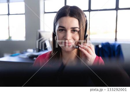 Woman in red blouse using headset microphone and speaking into computer monitor at desk near window 133292001