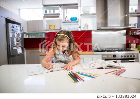 Girl child sitting at white countertop island in kitchen coloring workbook with colored pencils 133292024