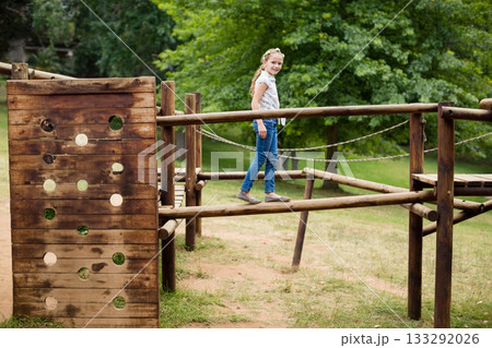 Female child balancing along wooden beam on park playground obstacle course with rope railings Female child balancing along wooden beam on park playground obstacle course with rope railings 133292026