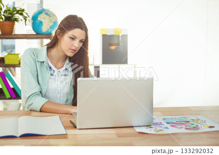 Woman sitting at desk working on laptop with notebook, color printouts in home office, copy space 133292032
