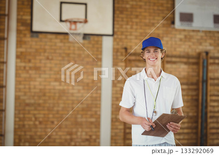 White teenage boy refereeing in gym holding clipboard, whistle on lanyard by hoop, copy space 133292069