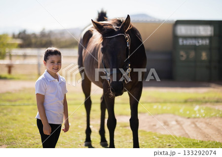 Smiling female child standing beside brown horse wearing halter in sunny paddock, copy space 133292074