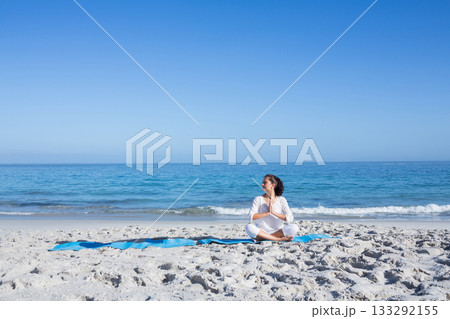 Woman practicing yoga seated on bright blue mat at sandy beach wearing white workout clothes Woman practicing yoga seated on bright blue mat at sandy beach wearing white workout clothes 133292155