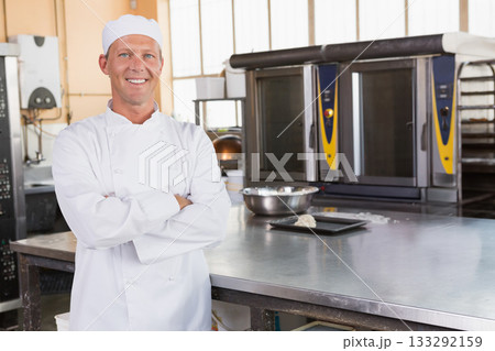 Senior male chef wearing jacket crossing arms in bakery beside steel worktable, copy space 133292159