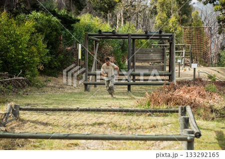 Male trainee sprinting through wooden hurdles and climbing rope net over mud patch on forest course 133292165