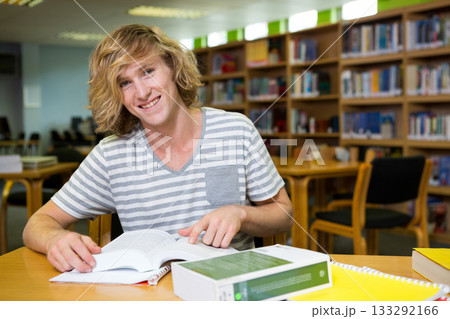 Male student studying open textbook, writing notes at library study table by green reference book 133292166
