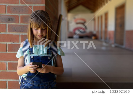 Female child using smartphone leaning on brick pillar in school hallway with backpack, copy space 133292169