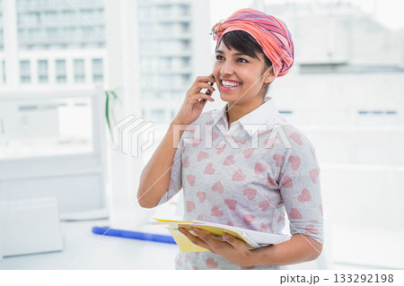 Asian woman standing by window in office wearing headscarf and holding notebook and smartphone 133292198