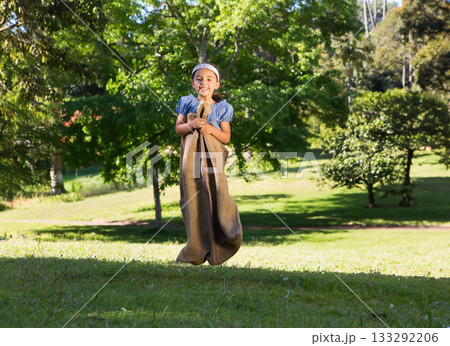 Boy jumping through burlap sack on lawn in sunshine wearing light blue top and white headband 133292206