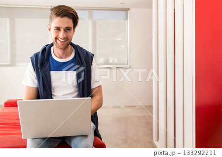 Male wearing navy sleeveless hoodie working on silver laptop on red bench in office lounge smiling 133292211