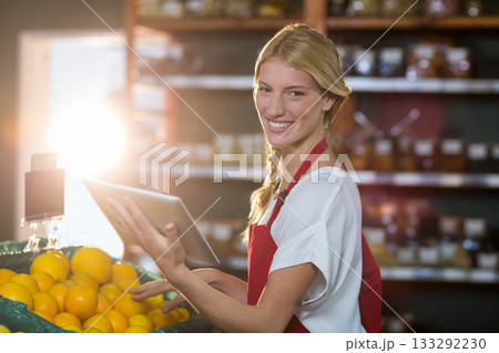 Female grocery clerk wearing red apron using tablet scanning oranges in produce section, copy space 133292230