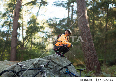 Female cyclist crouching on mossy rock in pine forest adjusting bike helmet beside mountain bikes 133292233