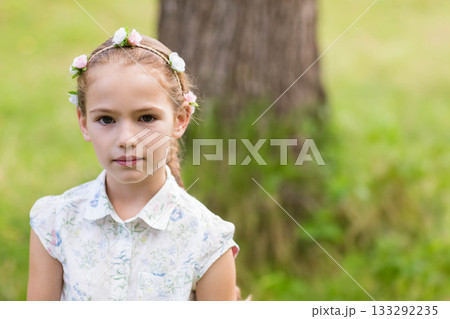 Female child standing beside tree trunk in meadow wearing floral headband and blouse, copy space Female child standing beside tree trunk in meadow wearing floral headband and blouse, copy space 133292235