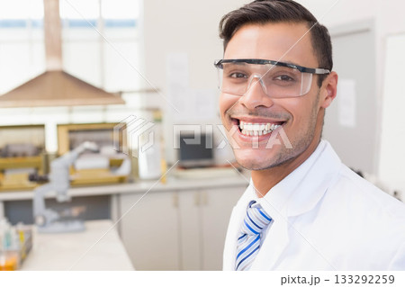 Man wearing lab coat, striped tie and safety goggles examining sample under microscope on lab bench 133292259