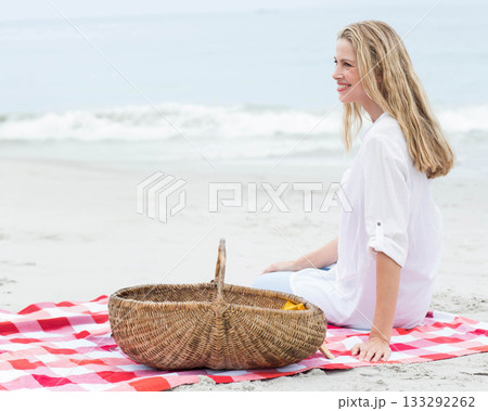 Senior woman sitting on checkered blanket on sandy shore beside wicker picnic basket, copy space 133292262
