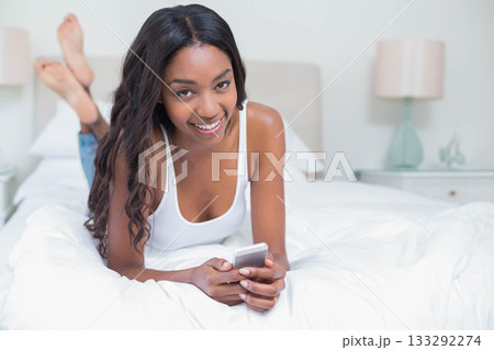 African American woman in her twenties lying on bed holding smartphone in softly lit bedroom 133292274
