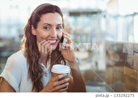 Woman holding disposable coffee cup while talking on smartphone at cafe table with headband 133292275