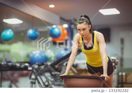 Female leaning forward on BOSU balance trainer in gym near wall-mounted stability balls, copy space 133292282