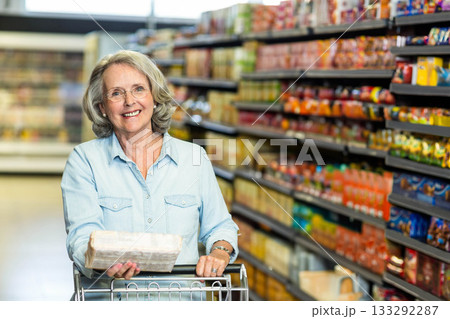 Senior woman pushing shopping cart and holding packaged loaf of sliced bread in supermarket aisle Senior woman pushing shopping cart and holding packaged loaf of sliced bread in supermarket aisle 133292287