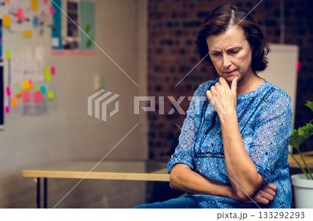 Senior female wearing blue blouse and jeans thinking at office table with planning board and plant 133292293