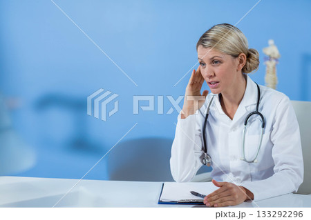 Female doctor sitting at desk in exam room reviewing clipboard beside spine model, copy space 133292296