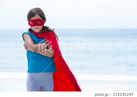 Female child striking superhero pose and standing on sandy shore wearing red cape and mask 133292297