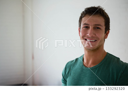 Man in twenties standing at home near vertical blinds wearing green shirt beside sofa, copy space Man in twenties standing at home near vertical blinds wearing green shirt beside sofa, copy space 133292320