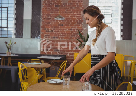 Female server wearing apron arranging clear glasses, plate, cutlery on cafe table, copy space Female server wearing apron arranging clear glasses, plate, cutlery on cafe table, copy space 133292322