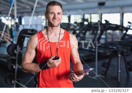 Mid adult trainer wearing athletic top holding stopwatch tablet in gym near weight benches, mirrors Mid adult trainer wearing athletic top holding stopwatch tablet in gym near weight benches, mirrors 133292327