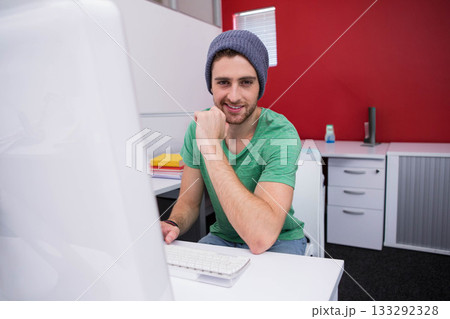 Male wearing green t-shirt and beanie sitting at desk in cubicle, leaning on fist beside notebooks 133292328