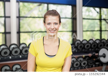 Standing in fitness center wearing yellow top and dark sports bra by dumbbell rack and dumbbells Standing in fitness center wearing yellow top and dark sports bra by dumbbell rack and dumbbells 133292344