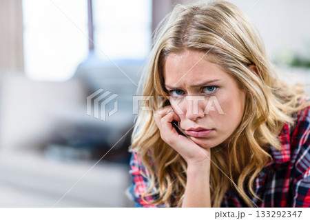 Woman sitting on sofa in living room wearing plaid shirt, resting chin on hand by window Woman sitting on sofa in living room wearing plaid shirt, resting chin on hand by window 133292347