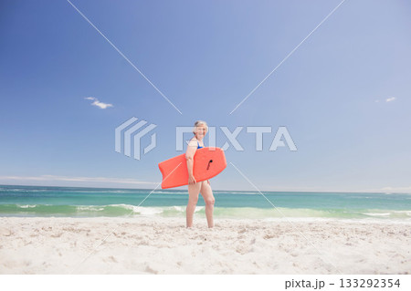 Senior woman standing on white-sand beach holding orange bodyboard, looking toward turquoise ocean 133292354