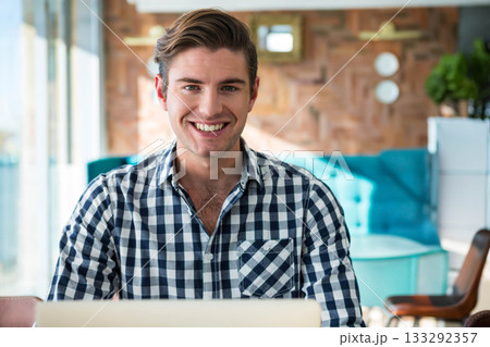 Male smiling while working on laptop in bright lounge area with turquoise sofa and plant Male smiling while working on laptop in bright lounge area with turquoise sofa and plant 133292357