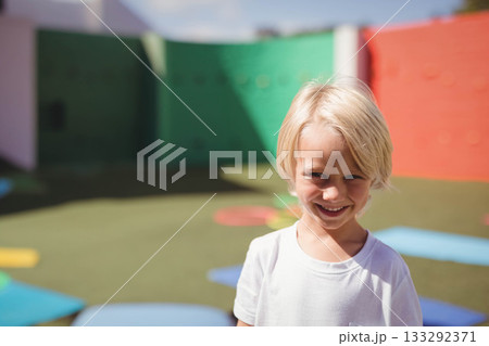 Smiling blond boy wearing white T-shirt on playground with foam mats, climbing holds, copy space 133292371