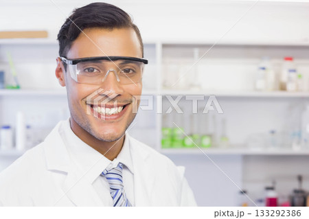 Smiling male scientist in safety goggles, lab coat, striped tie posing in lab with green solution Smiling male scientist in safety goggles, lab coat, striped tie posing in lab with green solution 133292386