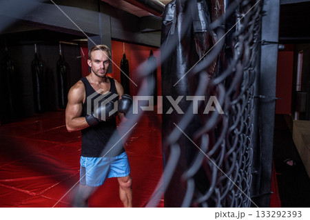 Male athlete training punches against heavy bag on red padded floor in gym with boxing gloves Male athlete training punches against heavy bag on red padded floor in gym with boxing gloves 133292393