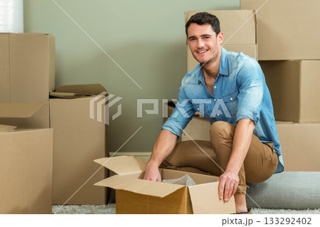 Man kneeling on carpet unpacking bubble wrap from moving box in new home wearing casual attire 133292402