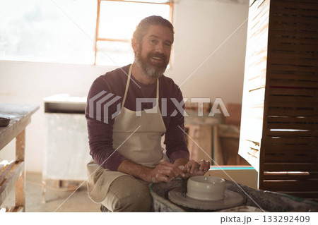 Middle-aged man wearing apron shaping wet clay cylinder on pottery wheel under window in workshop Middle-aged man wearing apron shaping wet clay cylinder on pottery wheel under window in workshop 133292409