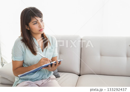 Asian woman in her twenties sitting on light sofa at home writing on clipboard, copy space Asian woman in her twenties sitting on light sofa at home writing on clipboard, copy space 133292437