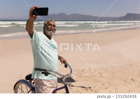 Senior Asian man standing on sandy beach beside bicycle holding smartphone taking selfie 133292438
