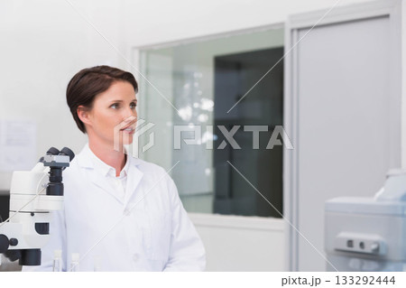 Female scientist wearing lab coat operating microscope in clinical lab beside diagnostic analyzer 133292444