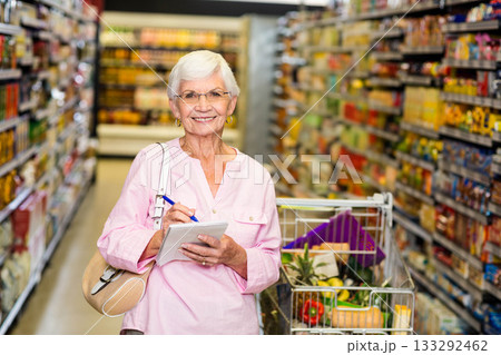Senior woman standing in supermarket aisle holding notepad with pen beside full cart with produce Senior woman standing in supermarket aisle holding notepad with pen beside full cart with produce 133292462